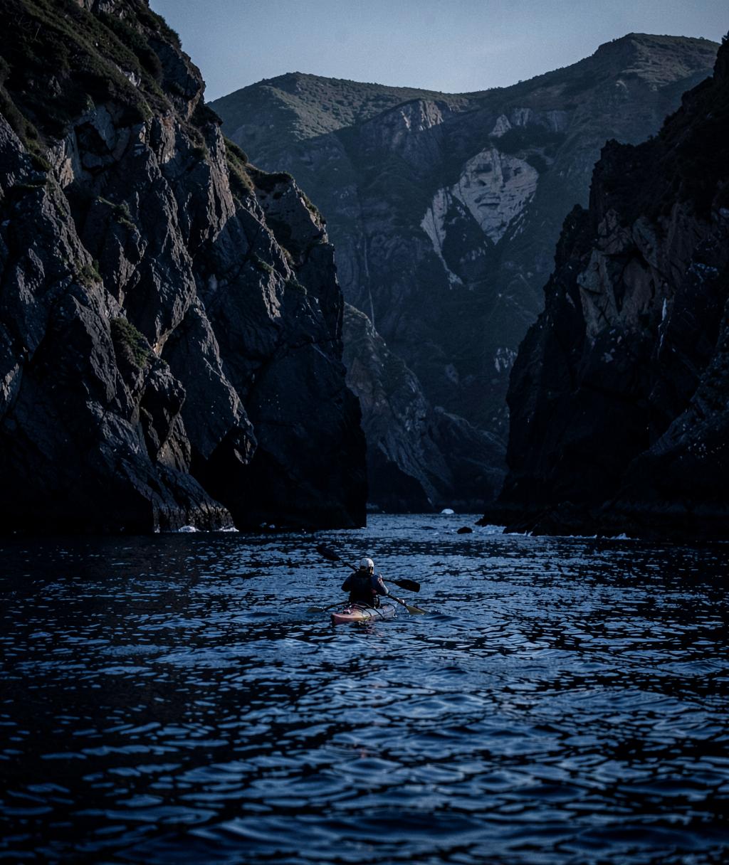 Lone sea kayaker paddling beneath rugged coastal cliffs at dawn